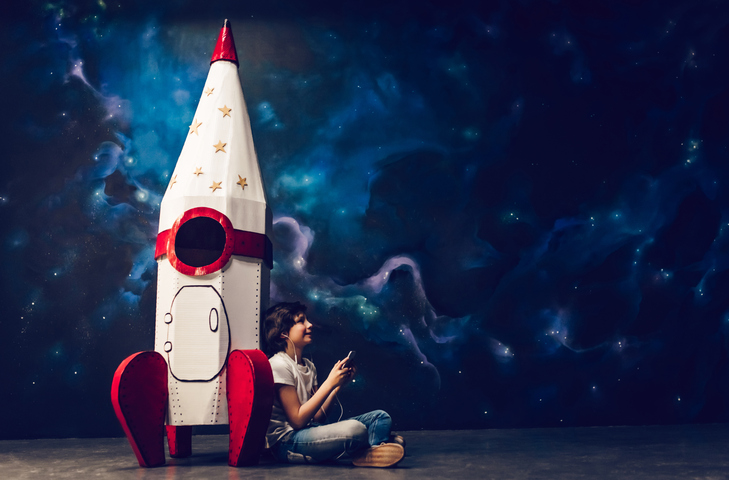 Young boy listening to music, sitting down and leaning against a big toy rocket ship.