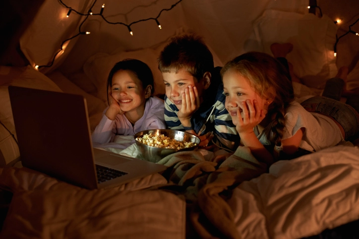 Happy kids in a tent watching a movie on a laptop while eating popcorn